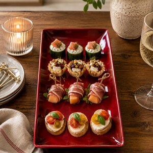 May include: A rectangular glossy red ceramic serving platter holding four rows of assorted appetizers, including cucumber rounds with salmon, pastry shells with cheese, prosciutto-wrapped melon, and crostini topped with tomatoes and basil.