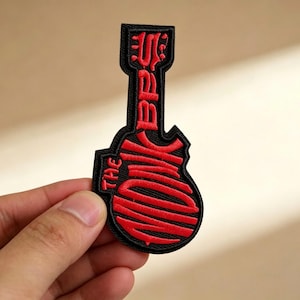 May include: Black and red embroidered patch in the shape of a guitar. The patch features the text "THE BACK" in red, stylized lettering. The patch is held by a hand against a neutral background.