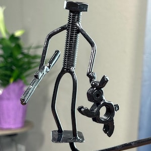 May include: A handcrafted metal sculpture depicting a figure holding a smaller figure. The larger figure is made from a bolt and other metal pieces, with arms and legs. The smaller figure appears to be a stylized animal. The sculpture is dark gray.