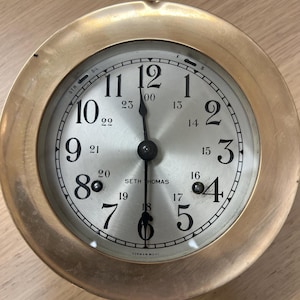 May include: A round brass clock with a silver face featuring black Arabic numerals from 1 to 12 and smaller 13 to 23 markers. The dial displays the text SETH THOMAS and GERMAN MOVI. It has three black hands and a small switch at the top labelled STR and SIL.