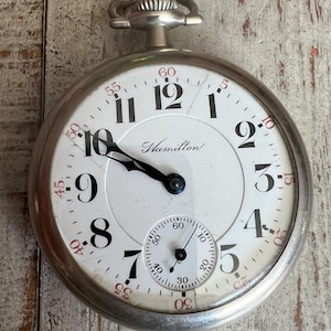 May include: A silver-tone Hamilton pocket watch with a white dial, black Arabic numerals, and black hands. The face features a smaller sub-dial for seconds at the bottom and red minute markers around the outer edge. A winding crown is at the top.