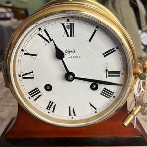 Pode incluir: Um relógio Schatz Royal Mariner com moldura circular de latão e mostrador branco com algarismos romanos pretos. O relógio tem ponteiros pretos, um ícone de sino perto da posição das três horas e uma chave de corda de latão na lateral.