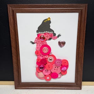 May include: A framed wall art piece featuring a silhouette of a figure wearing a gown constructed from various pink buttons, beads, and gems. The figure has a dark hair silhouette, a pearl-like necklace, and a small gold crown, with a pink heart.