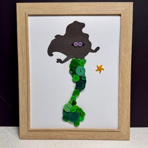 May include: A framed wall art piece featuring a dark silhouette of a mermaid figure. The tail is constructed from various shades of green buttons in different sizes. Two small purple buttons form the eyes, and a yellow starfish is placed nearby.