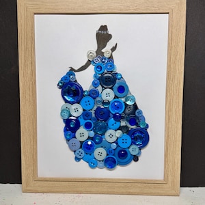 May include: A framed artwork featuring a silhouette of a figure wearing a voluminous gown constructed from a collage of various blue buttons in shades of navy, royal, and light blue, accented with clear faceted gems on a white background.