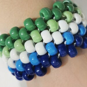 Puede incluir: Una pulsera de cuentas multicolores. La pulsera presenta un degradado de cuentas verdes, blancas y azules. Las cuentas están ensartadas juntas, creando un efecto de capas. La pulsera está hecha de cuentas de diferentes tamaños.