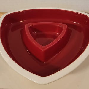May include: A triangular ceramic serving dish featuring a deep red interior and a contrasting white outer rim. The design includes a smaller, raised triangular compartment centered within the larger triangular basin, creating a two-section layout.
