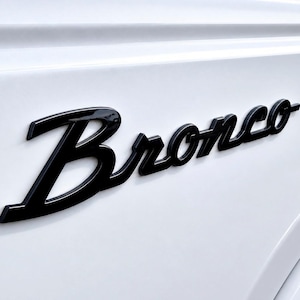 Peut inclure: Gros plan sur la carrosserie blanche d'un véhicule avec l'emblème "Bronco" noir brillant. L'emblème est en écriture cursive, mettant en valeur le design épuré et l'image de marque du véhicule.