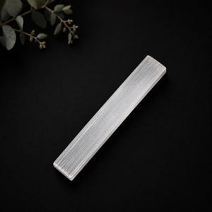 May include: A rectangular selenite crystal wand with a translucent, white, fibrous appearance and a smooth, polished surface. The crystal features distinct vertical striations running along its length, creating a linear, reflective texture.