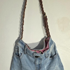 May include: A denim handbag made from repurposed jeans, featuring a brown braided strap and a fringed bottom. The bag has a light blue wash with distressed details and a floral patterned lining.