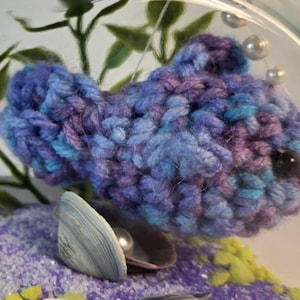Op de afbeelding: Een handgemaakte gehaakte vis in blauwe en paarse tinten, met een zwart kraaloog. De vis wordt gepresenteerd in een glazen kom met decoratieve elementen, waaronder een zeeschelp, parels en paars zand. Een uniek, handgemaakt item.