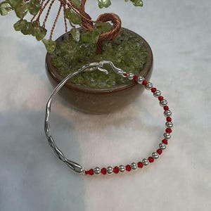 Puede incluir: Una pulsera de plata con un diseño orgánico único. Un lado es una forma sólida y fluida, mientras que el otro presenta pequeñas cuentas plateadas ensartadas en un cordón rojo. La pulsera está cerca de un pequeño bonsái con hojas verdes.