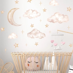 May include: A wall decal set featuring a crescent moon, fluffy clouds, and various stars in soft beige and pink tones. A wooden crib sits below with a hanging mobile featuring pink hearts and white stars, plus two plush pillows inside.