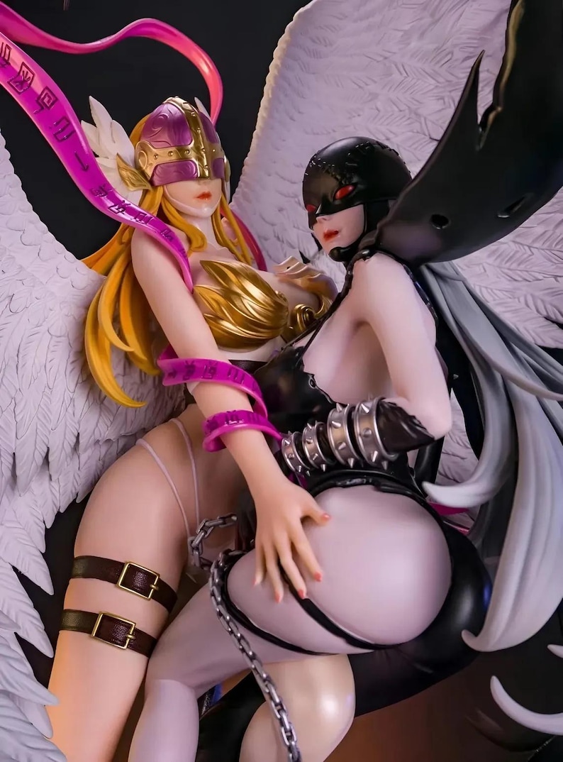 May include: A detailed figurine featuring two female characters with elaborate costumes and wings. One figure has blonde hair, a gold breastplate, and a pink headpiece. The other figure is in black attire with a mask. The wings are white and black.