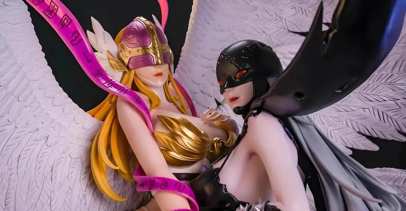 May include: A detailed figurine featuring two female characters with angelic and demonic themes. One figure has blonde hair, a gold and purple helmet, and white wings. The other has black wings and a black helmet. Both figures have detailed armour.