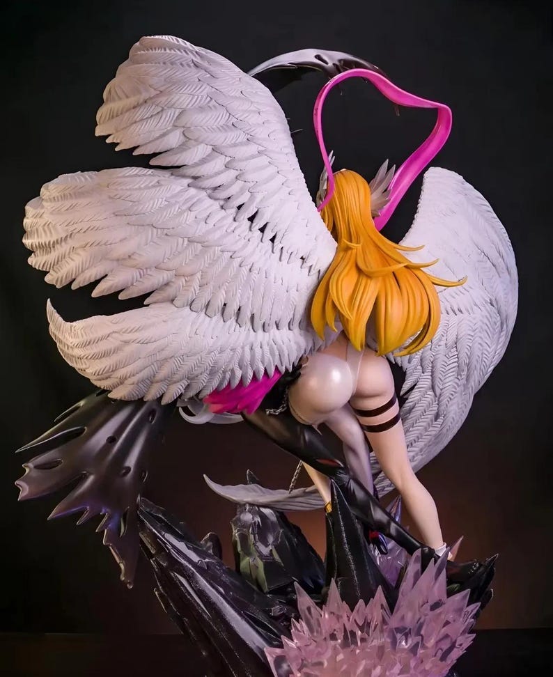 May include: A detailed figurine of a woman with long, flowing blonde hair, wearing a white and black outfit, and large white feathered wings. The figure is posed with a pink heart-shaped accent and stands on a dark, textured base with crystal-like elements.
