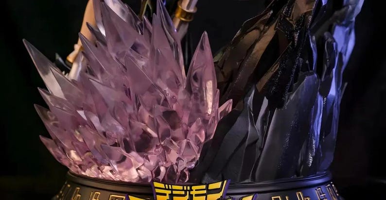 May include: A detailed statue base featuring a central cluster of translucent, purple crystal formations. The base has a black and gold trim with Japanese text. The background is dark, highlighting the statue's intricate design.