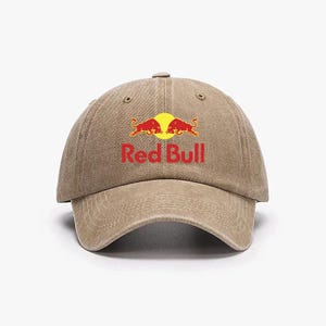 Könnte beinhalten: Beige Baseball Cap mit dem Red Bull Logo. Das Logo zeigt zwei rote Bullen, die sich vor einer gelben Sonne gegenüberstehen, darunter die roten Worte "Red Bull". Die Kappe hat einen gebogenen Schirm und einen Stoffriemen.