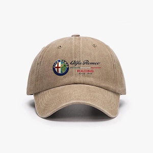 Può includere: Un cappellino da baseball beige con il logo e il testo Alfa Romeo. Il logo è un emblema circolare con una croce rossa e un serpente verde. Il testo "Alfa Romeo Racing Since 1910" è visualizzato in nero e rosso.