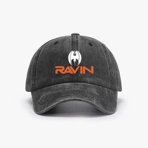 May include: A black baseball cap with the word "RAVIN" in orange block letters. Above the word is a white graphic of a stylized bird or bat. The cap has a curved brim and a fabric strap closure.