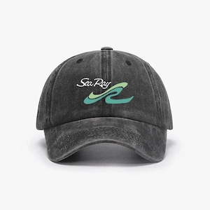 Op de afbeelding: Een zwarte, verwassen baseballcap met de tekst "Sea Ray" in witte letters boven een gestileerd golfontwerp in teal en groen. De cap heeft een gebogen klep en een knoop aan de bovenkant.