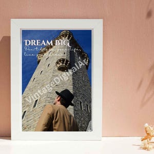 Puede incluir: Una impresión artística enmarcada en blanco con una torre de piedra bajo un cielo azul. Una persona con chaqueta beige y sombrero negro mira hacia la torre. El texto dice: DREAM BIG. Don't doze in your life. Live your dream. VintageDigitalArt es visible.