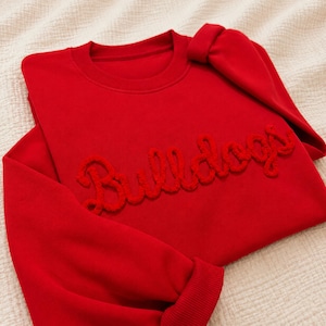 May include: A bright red crewneck sweatshirt featuring the word Bulldogs written in a raised, textured, cursive script across the chest. The garment has long sleeves with ribbed cuffs and a matching ribbed neckline, all in a solid red color.