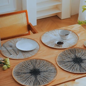 Könnte beinhalten: Ein Holztisch mit vier runden Tischsets mit einem radialen Design in Weiß und Schwarz. Jedes Tischset hat einen hellbraunen Rand. Weiße Teller und kleine Schalen sind auf einigen Tischsets platziert.