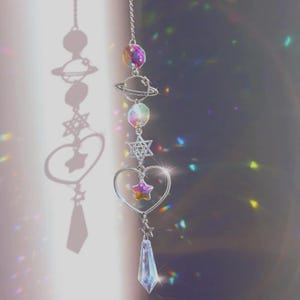 May include: A celestial-themed suncatcher with a chain, featuring iridescent crystal planets, stars, and a heart. The design includes a large crystal pendant at the bottom, casting a shadow of the same shapes. The colors are primarily silver and rainbow.