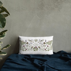 May include: A rectangular white pillow with a nature-themed design. The design features green moths, dragonflies, and various plants and flowers in shades of green, purple, and brown. The pillow is placed on a dark blue blanket.