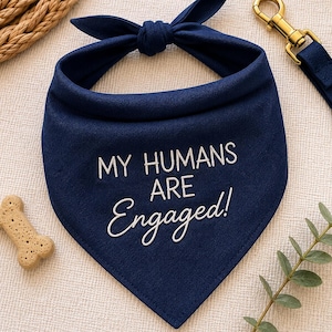 May include: A navy blue dog bandana with the text "MY HUMANS ARE Engaged!" in white script. A gold-coloured leash clip and a bone-shaped dog treat are also visible.
