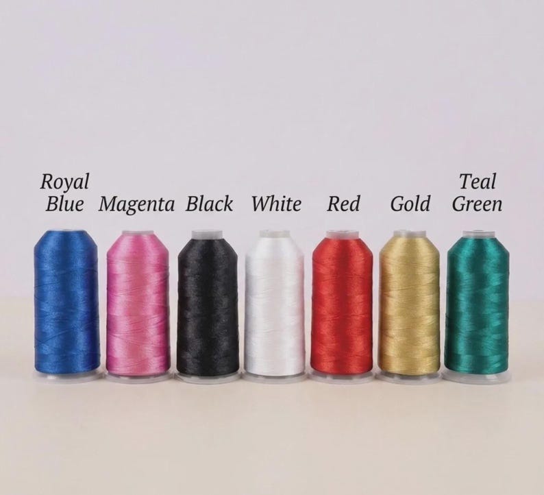 May include: Seven spools of embroidery thread in a row, with colours including royal blue, magenta, black, white, red, gold, and teal green. Each spool is labelled with its colour name. The background is white.