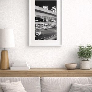 May include: Monochrome framed photograph of a city street at night, showing a highway overpass with vehicles and illuminated signs. The photograph is displayed on a wooden surface with a lamp, books, and a potted plant.
