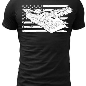 May include: Black t-shirt featuring a white graphic of a futuristic vehicle design set against a stylized American flag. The design includes stars and stripes, with the vehicle appearing to be a type of spacecraft or military transport.