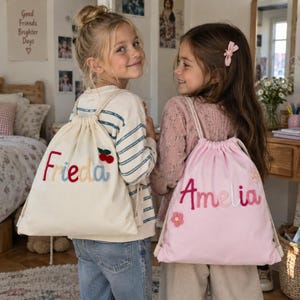May include: Two drawstring backpacks made of fabric. The off-white bag features the name Frieda embroidered in multi-colored letters with a small red cherry applique. The light pink bag features the name Amelia embroidered in pink tones with small flower appliques.