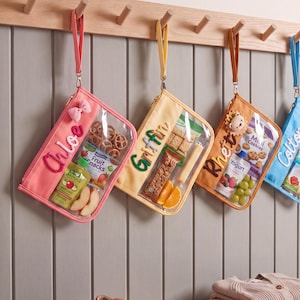 Pode incluir: Quatro bolsas de pulso retangulares com frente de vinil transparente e verso de tecido de cor sólida em rosa, amarelo, laranja e azul. Cada bolsa apresenta um nome personalizado em letras de feltro, uma alça de pulso e contém vários lanches.