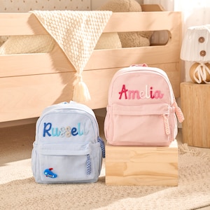Peut inclure: Un sac à dos en velours côtelé bleu clair avec le prénom Russell et un petit écusson d'avion, et un sac à dos en velours côtelé rose avec le prénom Amelia brodé en dégradé rose. Les deux sacs ont des poches avant zippées à tirettes tressées.