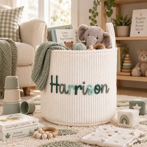 May include: A cream-coloured, ribbed fabric storage basket featuring the name Harrison embroidered in dark green and light blue yarn. The basket is filled with a grey plush elephant, a book titled Little Bear Big Dreams, and a sage green knit blanket.