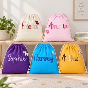 May include: Five drawstring backpacks in cream, pink, purple, blue, and yellow, each featuring a personalized name embroidered in cursive script and a small decorative felt applique, such as a cherry, flower, unicorn, whale, or lion.