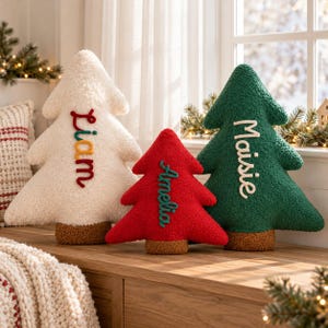 May include: Three tree-shaped plush pillows with textured fabric. The white tree features the name Liam in red, orange, and green. The red tree features the name Amelia in green. The green tree features the name Maisie in white.