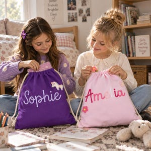 May include: Two drawstring backpacks made of fabric, one purple with the name Sophie and a unicorn patch, and one light pink with the name Amelia and flower patches. Both bags feature thick white rope straps and personalized lettering.