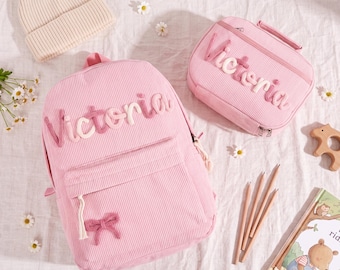 May include: A light pink corduroy backpack and a matching rectangular pencil case, both featuring the name Victoria embroidered in cursive script. The backpack includes a small pink bow detail on the front pocket, accompanied by a beige knit beanie.