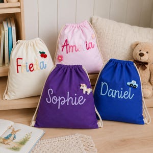 Pode incluir: Quatro mochilas de cordão em creme, rosa claro, roxo e azul. Cada bolsa apresenta um nome personalizado em letras de feltro: Frieda com cerejas, Amelia com flores, Sophie com um unicórnio e Daniel com um carro. As bolsas têm cordas grossas.