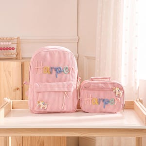 May include: A pink corduroy backpack and a matching rectangular lunch bag, both featuring the name Harper in colorful, raised, multi-textured letters. Each item is decorated with a small, embroidered unicorn patch with a gold horn.