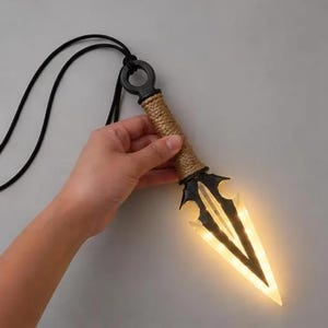 May include: A kunai-shaped pendant necklace featuring a black ring at the top, a textured tan handle, and a translucent, glowing blade that emits a warm light. The pendant is attached to a thin black cord, creating a functional accessory piece.