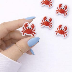 May include: Embroidered crab patches in red and white, with black eyes and a white border. The patches are small, perfect for adding to clothing or accessories. The image shows several patches on a white background.