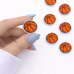 May include: Embroidered basketball patches with orange and black detailing. The patches are round with a white border. One patch is held by a hand with blue nail polish, and several others are scattered on a white surface.