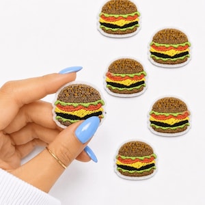 May include: Five small embroidered hamburger patches featuring a brown bun, green lettuce, red tomato, yellow cheese, and a black patty. Each patch has a white border and is circular in shape, showcasing detailed stitching of a classic cheeseburger.