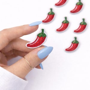 May include: Embroidered red chili pepper patches with green stems. One patch is held between fingers with light blue nail polish. Several other patches are scattered on a white background. The patches are a fun, decorative item.