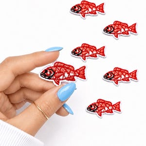 May include: Embroidered red fish patches with black details and white borders are scattered on a white background. The patches are in various orientations. One patch is held by a hand with a gold ring and blue nail polish.